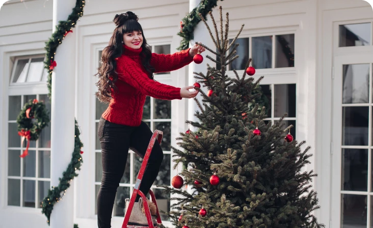 Femeie decorând un brad de Crăciun într-un spațiu decorat festiv, îmbrăcată într-un pulover roșu, atârnând globuri roșii în brad.