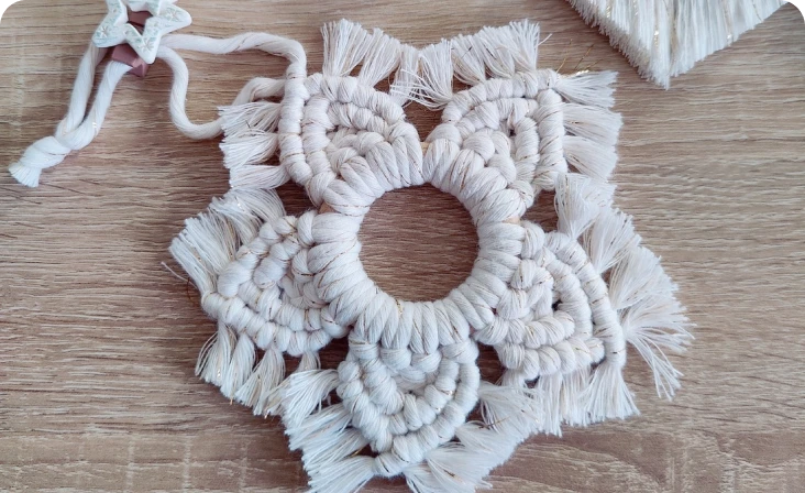 Ornament de Crăciun realizat manual în formă de fulg de nea din macrame, așezat pe o suprafață de lemn.